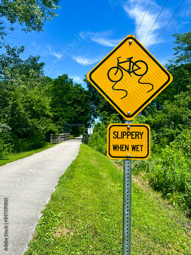 "Slippery When Wet" Yellow Warning sign located beside a bicycle path. Rural setting.