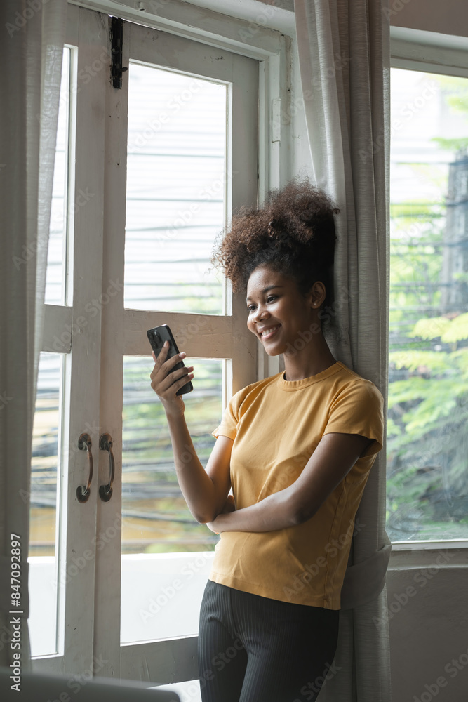 Young African Woman Smiling While Using Smartphone at Home by the Window, Casual Clothing, Natural Light, Modern Lifestyle, Relaxed and Happy, Indoor Setting, Daytime, Technology and Communication Young African Woman Smiling While Using Smartphone at Home by the Window, Casual Clothing, Natural Light, Modern Lifestyle, Relaxed and Happy, Indoor Setting, Daytime, Technology and Communication