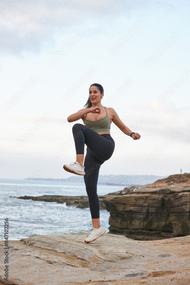 Female hispanic runner doing kick jumps, wearing black leggings and a olive top, ocean view in the background. Female hispanic runner doing kick jumps, wearing black leggings and a olive top, ocean view in the background.