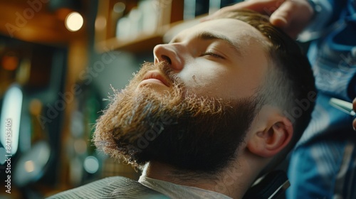 A man with a thick beard relaxes in a barber shop as a stylist prepares to trim his facial hair. He has his eyes closed and appears to be enjoying the experience.