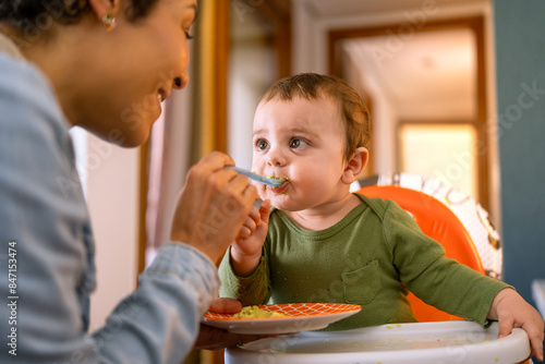 Loving mother spoon-feeding her happy baby boy. Adorable toddler in green outfit enjoying a nutritious meal at home..