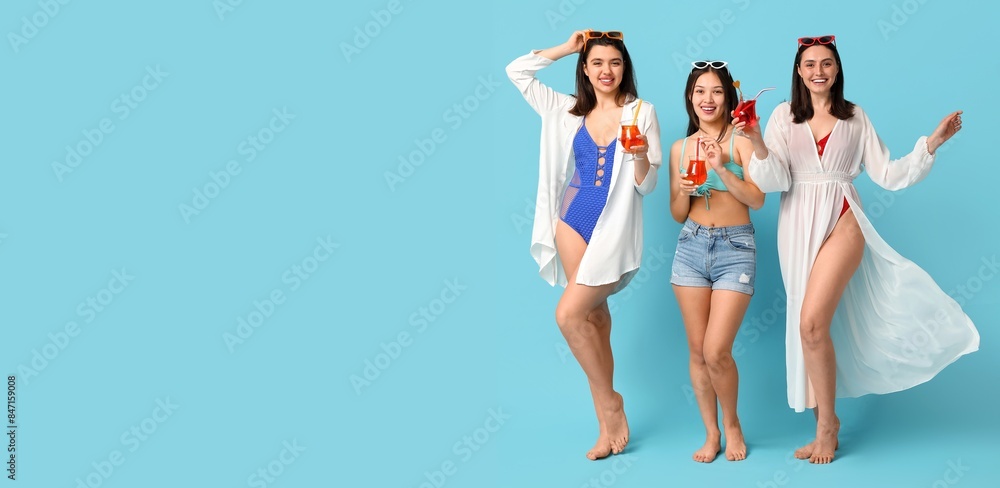 Obraz premium Happy young women in swimsuits and with glasses of cocktail on blue background with space for text