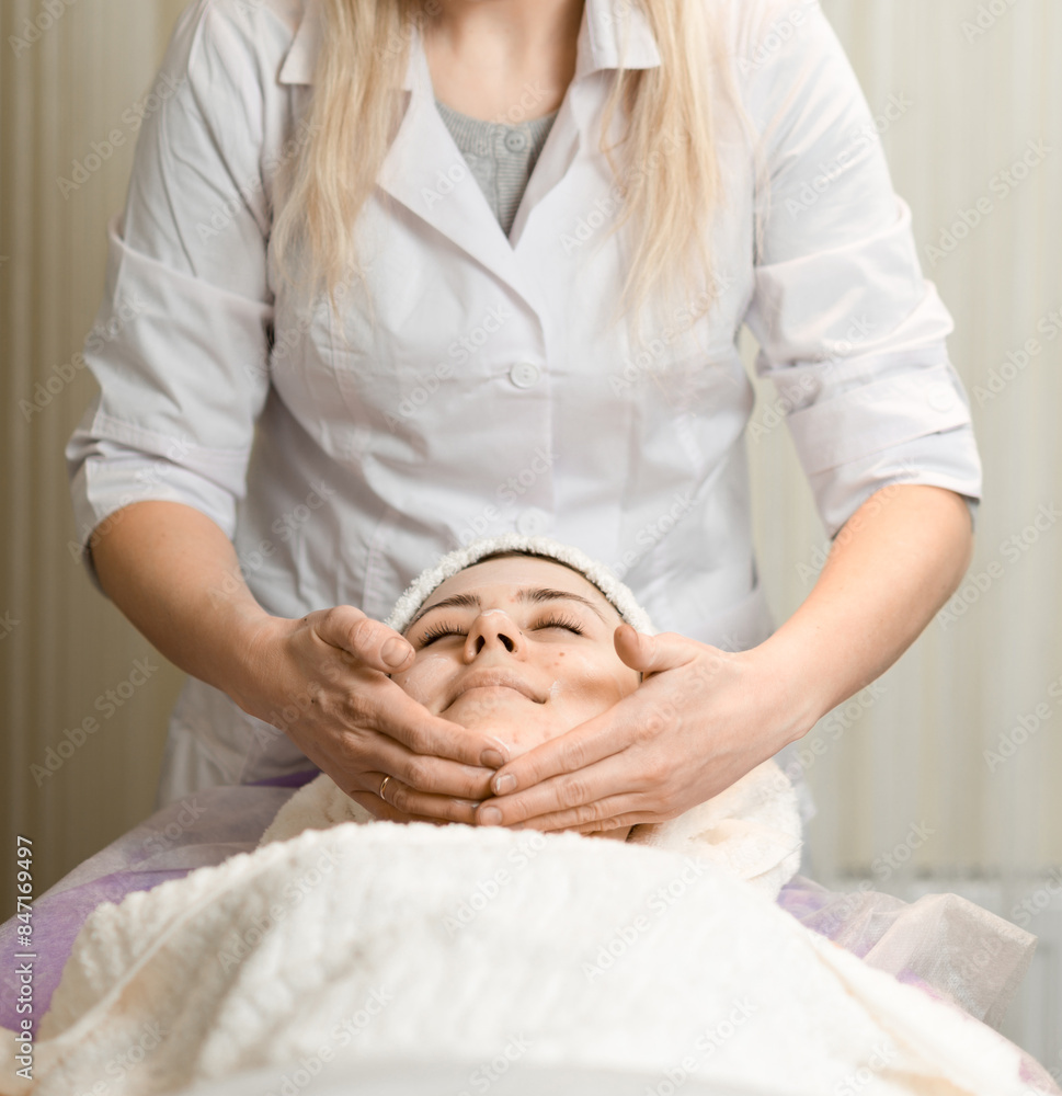 Obraz premium Young woman enjoying facial massage, lying on spa bed indoor at beauty salon.