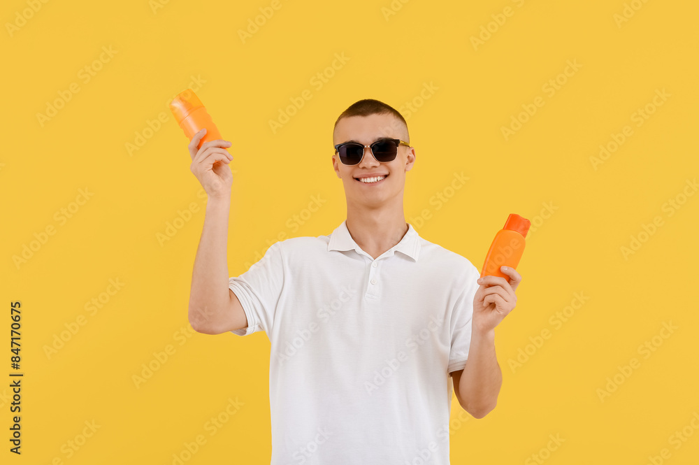 Happy young man in sunglasses with bottles of sunscreen cream on yellow background Happy young man in sunglasses with bottles of sunscreen cream on yellow background
