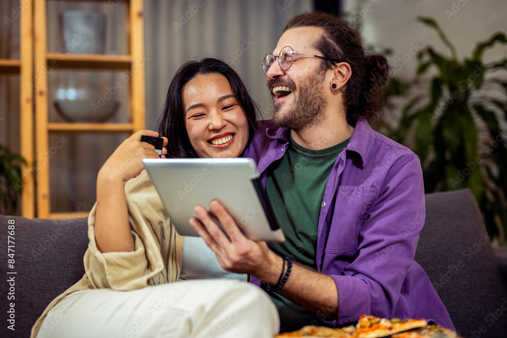 A couple, an Asian woman and a white man, is comfortably seated in their cozy living room. They are engaged in making an online payment together, A couple, an Asian woman and a white man, is comfortably seated in their cozy living room. They are engaged in making an online payment together,
