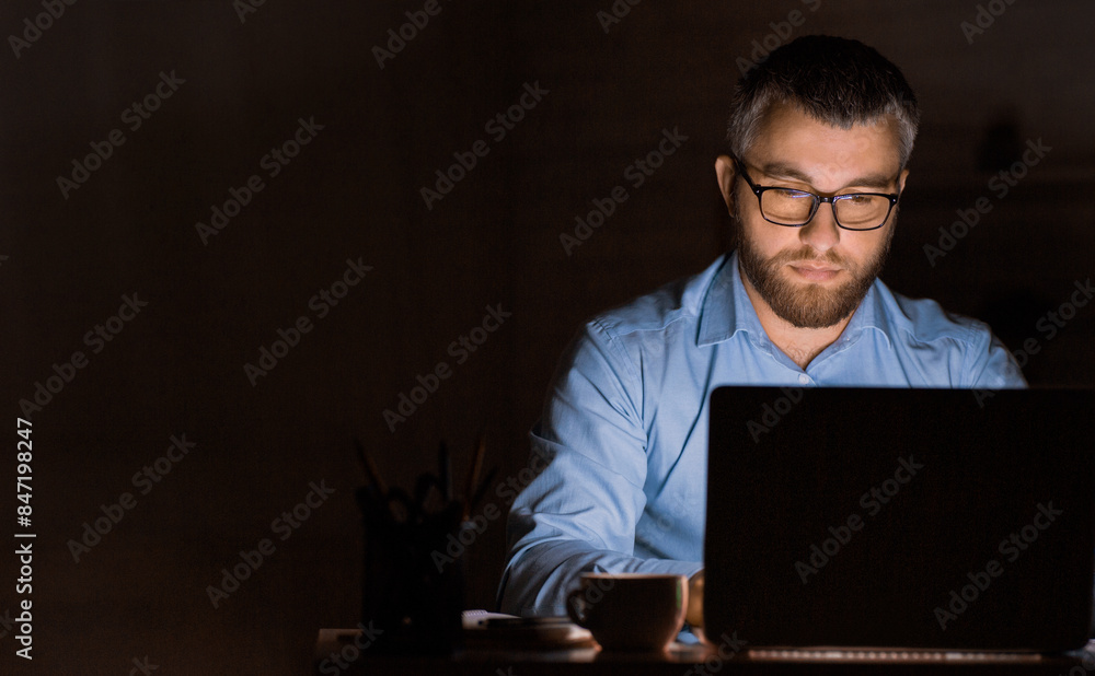 A man with a beard and glasses wearing a blue shirt is focused on his laptop, working late at night in a dimly lit home office. A man with a beard and glasses wearing a blue shirt is focused on his laptop, working late at night in a dimly lit home office.