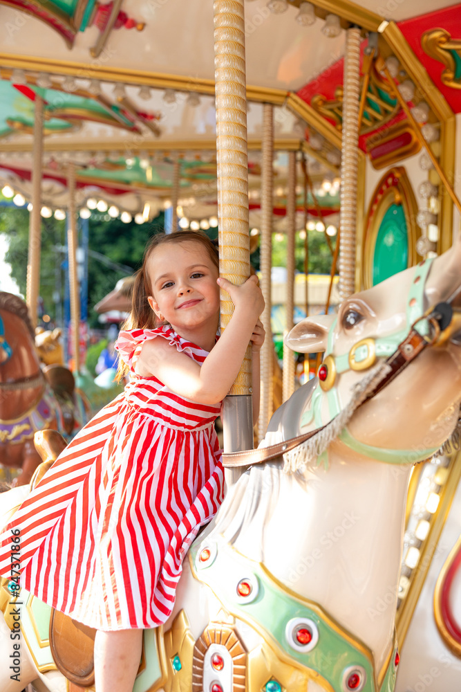 Obraz premium Adorable little brunette girl in summer red-white dress at amusement park having a ride on the merry-go-round. Child girl has fun outdoor on sunny summer day. Entertainment concept