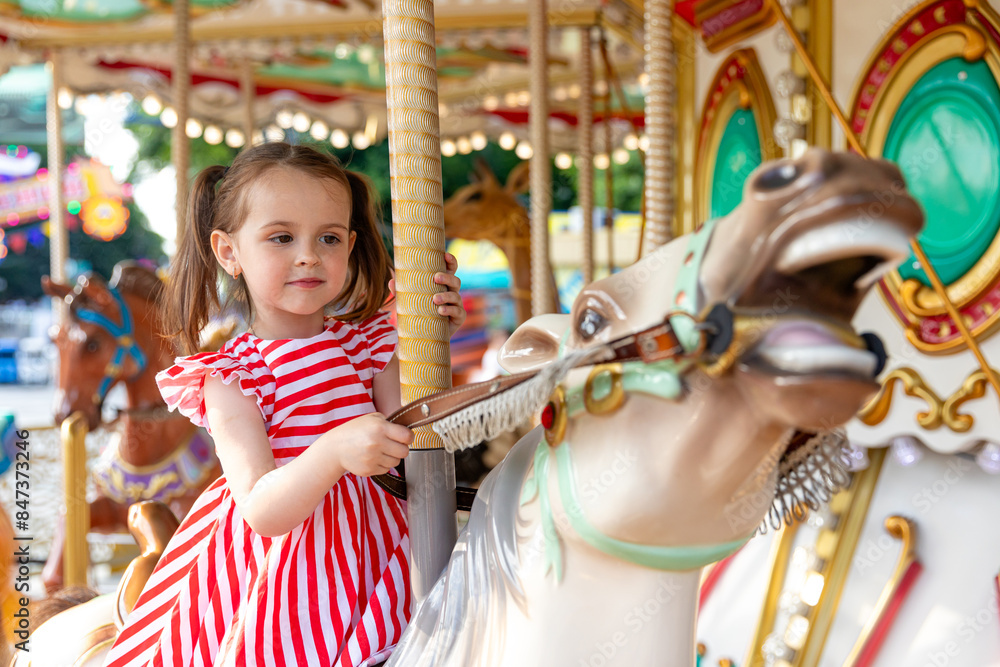 Adorable little brunette girl in summer red-white dress at amusement park having a ride on the merry-go-round. Child girl has fun outdoor on sunny summer day. Entertainment concept