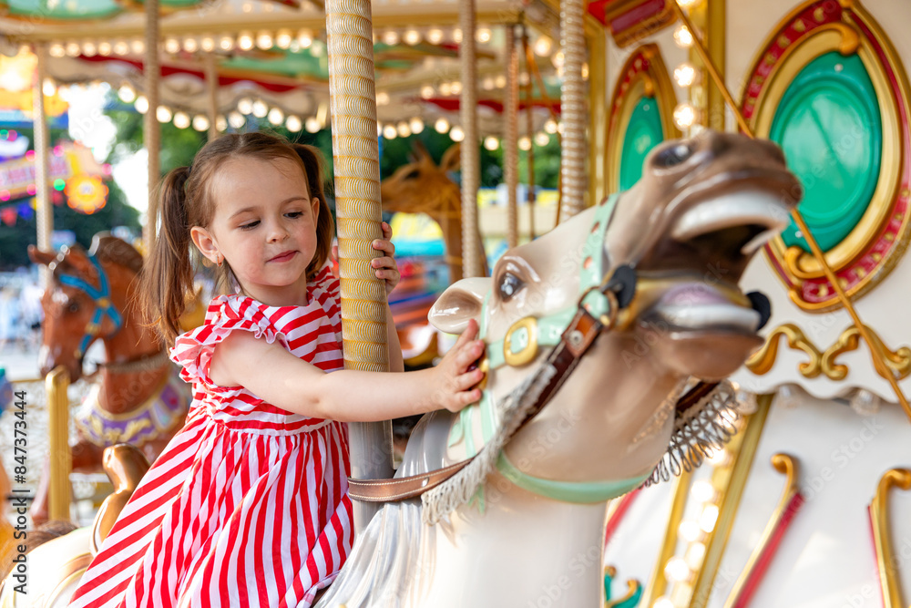 Fototapeta premium Adorable little brunette girl in summer red-white dress at amusement park having a ride on the merry-go-round. Child girl has fun outdoor on sunny summer day. Entertainment concept