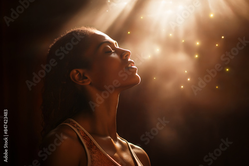 Woman of faith with eyes closed in prayer is blessed with divine light shining on her face. She looks full of devotion & grace, experiencing the bliss of a mystical experience, a spiritual awakening.