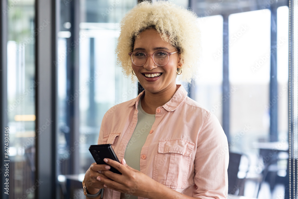 © Wavebreak Media - Young biracial woman with curly blonde hair holding smartphone and smiling in business office