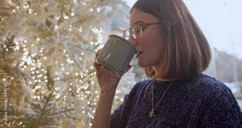 Indoors, by a beautifully decorated, illuminated Christmas tree, a young woman in a cozy sweater sips from a mug, embodying warmth and festive spirit, creating a cozy and joyful holiday atmosphere. photo