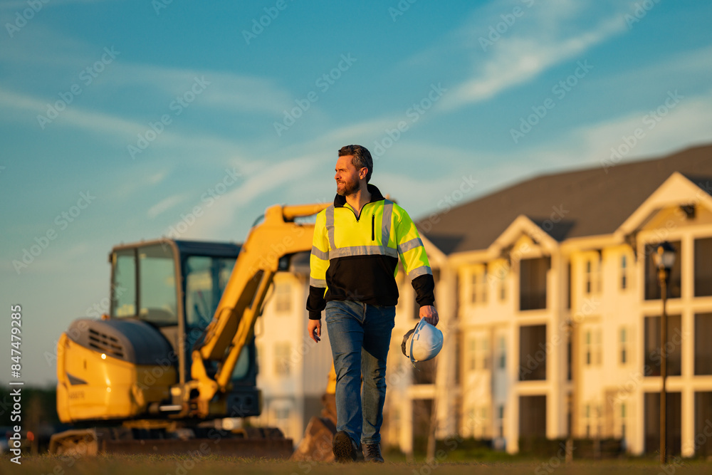 Builder in a construction site. Builder with excavator ready to build new house. Construction builder wear building uniform on excavation truck digging, builder with buildings construction background. Builder in a construction site. Builder with excavator ready to build new house. Construction builder wear building uniform on excavation truck digging, builder with buildings construction background.