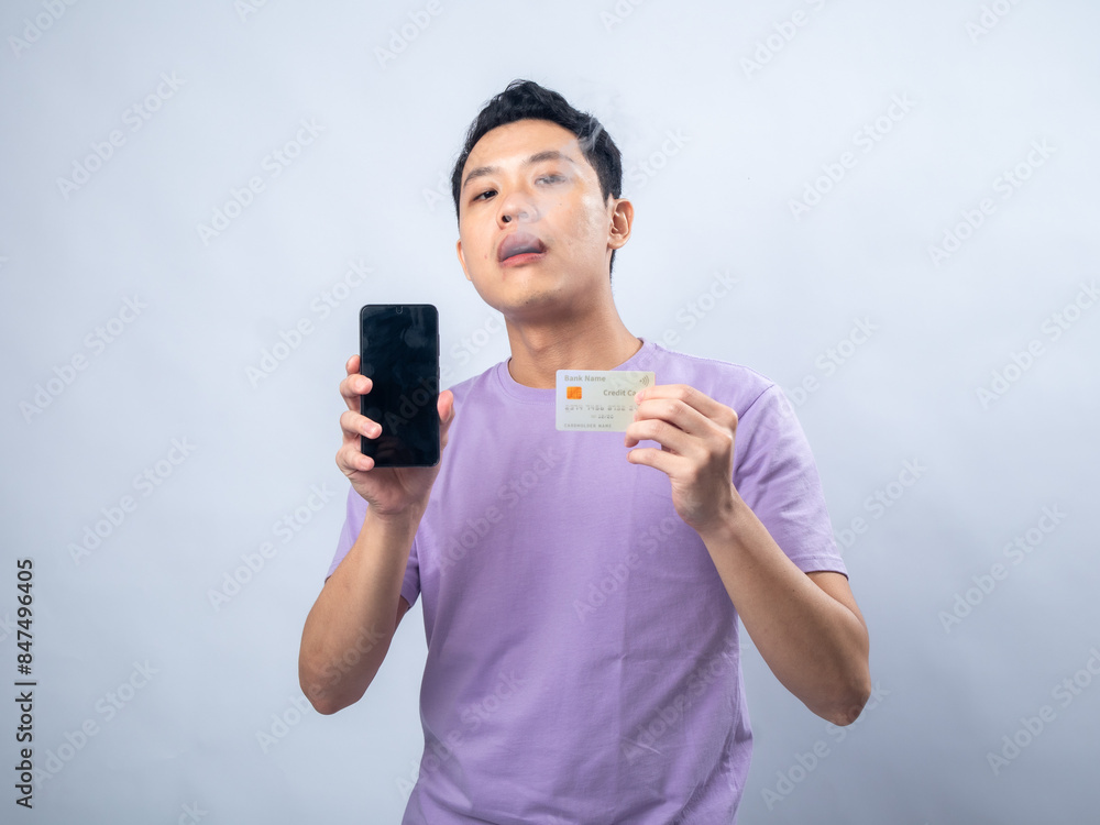 Smiling young man in a lavender shirt, holding a smartphone in one hand and a credit card in the other, against a plain light gray background. The man exudes confidence and modernity Smiling young man in a lavender shirt, holding a smartphone in one hand and a credit card in the other, against a plain light gray background. The man exudes confidence and modernity