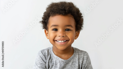 A close-up of a joyful young African-American boy with curly hair, beaming with a bright smile. Wearing a light grey t-shirt, his expressive eyes radiate happiness against a plain white background. © Kevin