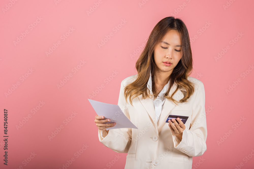 Portrait of Asian young woman holding paper calculating bills on hand and credit card for payment, female person problem she hold worried over bill, studio shot isolated on pink background Portrait of Asian young woman holding paper calculating bills on hand and credit card for payment, female person problem she hold worried over bill, studio shot isolated on pink background