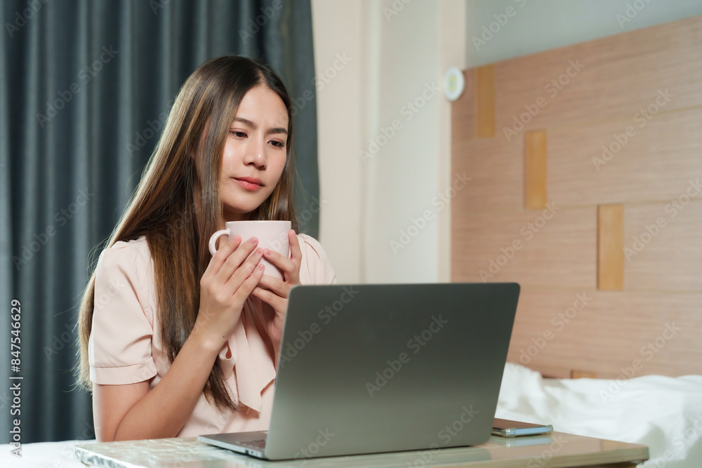 Beautiful Asian woman holding cup while working on laptop. Long straight hair, focused expression, casual clothing. Indoor home office setting with curtains and wooden wall in background. Beautiful Asian woman holding cup while working on laptop. Long straight hair, focused expression, casual clothing. Indoor home office setting with curtains and wooden wall in background.