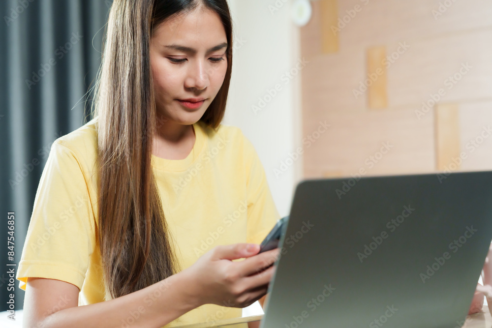 Asian woman in yellow shirt working on laptop, using smartphone. Indoor home office with wooden wall. Focused expression, casual attire, modern workspace, long straight hair, digital communication. Asian woman in yellow shirt working on laptop, using smartphone. Indoor home office with wooden wall. Focused expression, casual attire, modern workspace, long straight hair, digital communication.