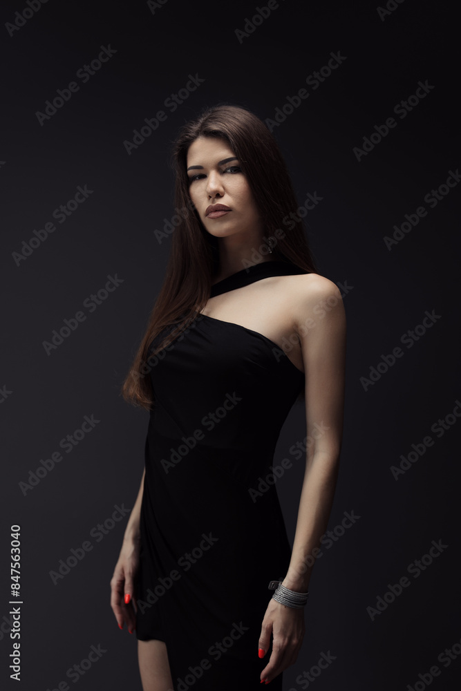 A poised young woman in a stylish black dress stands against a dark backdrop, exuding elegance and confidence with a hint of mystery. Perfect for fashion and lifestyle themes.