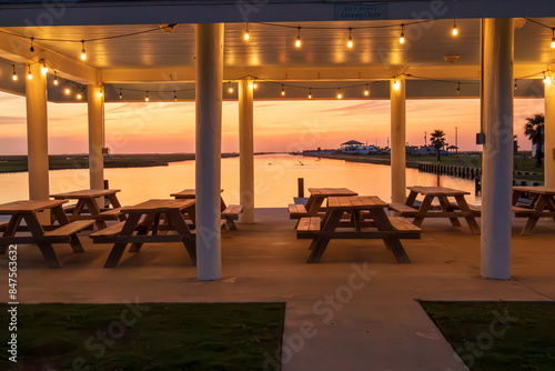 Canvas Print Picnic tables are neatly arranged under a covered pavilion adorned with hanging lights, providing a prime spot to enjoy the serene sunset over the calm waters in Freeport Texas