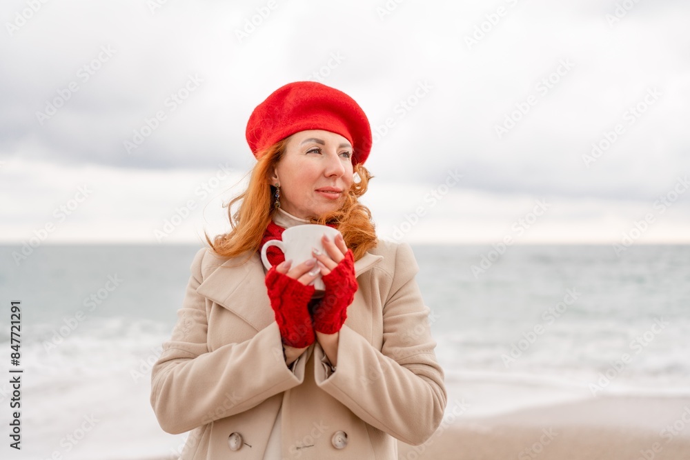 Obraz premium Woman beach in red beret scarf and mitts holding a cup of tea in his hands. Depicting beach relaxation and cozy attire. Walks by the sea