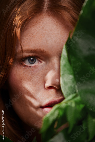 Beauty portrait of a young woman with red hair, flawless skin and freckles, surrounded by tropical leaves, like in the jungle. A feeling of curiosity. 