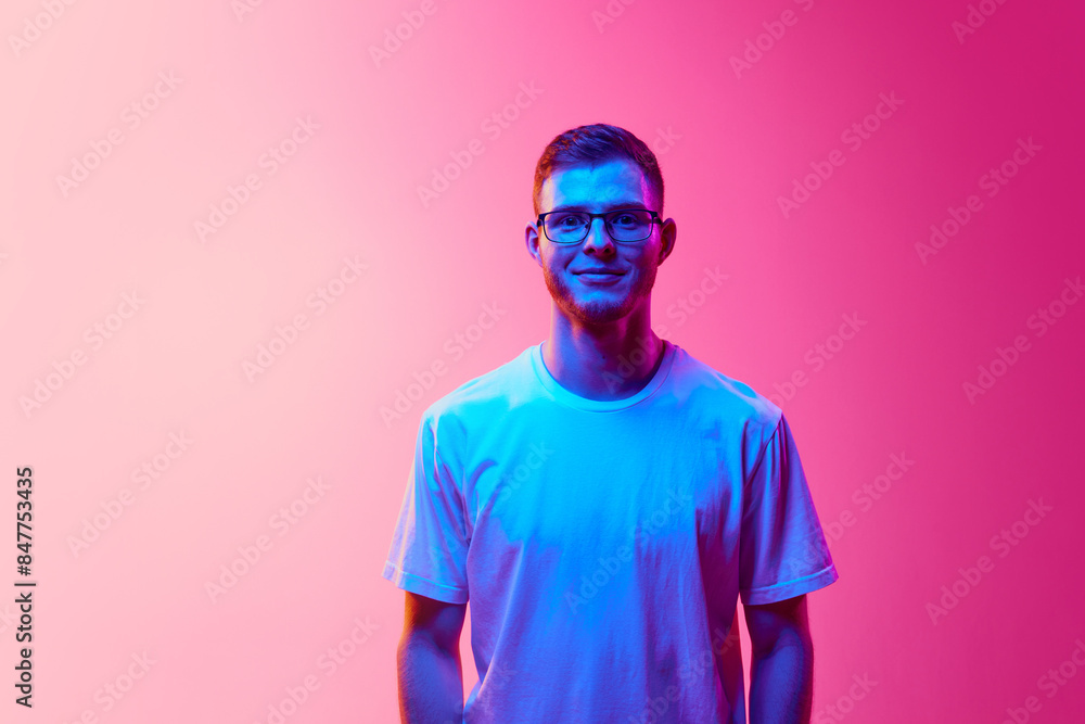 Portrait of young man in casual white t-shirt and glasses posing, looking at camera against pink background in neon light. Concept of human emotions, facial expression, lifestyle Portrait of young man in casual white t-shirt and glasses posing, looking at camera against pink background in neon light. Concept of human emotions, facial expression, lifestyle