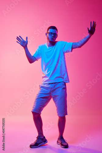 Full-length image of young man in glasses and casual clothes cheerfully dancing and having fun against pink background in neon light. Concept of human emotions, facial expression, lifestyle © master1305