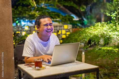 Young Asian man, digital nomad working remotely from cafe, using laptop and smartphone. young Asian men are hanging out in a cafe with a nature theme at night