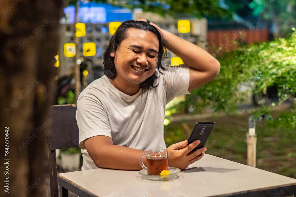 Young Asian man, digital nomad working remotely from cafe, using laptop and smartphone. young Asian men are hanging out in a cafe with a nature theme at night