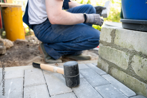 Worker in coverall laying paving stones with rubber mallet on house backyard