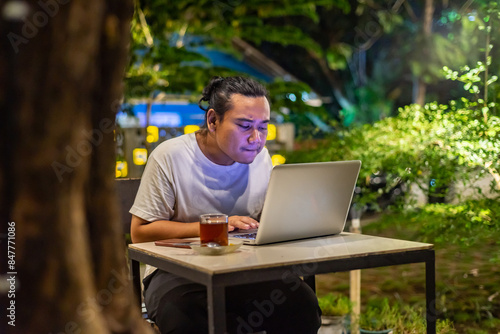Young Asian man, digital nomad working remotely from cafe, using laptop and smartphone. young Asian men are hanging out in a cafe with a nature theme at night