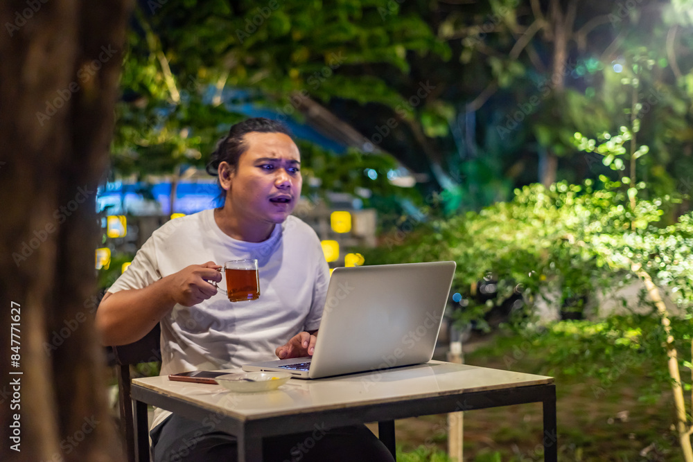 Young Asian man, digital nomad working remotely from cafe, using laptop and smartphone. young Asian men are hanging out in a cafe with a nature theme at night