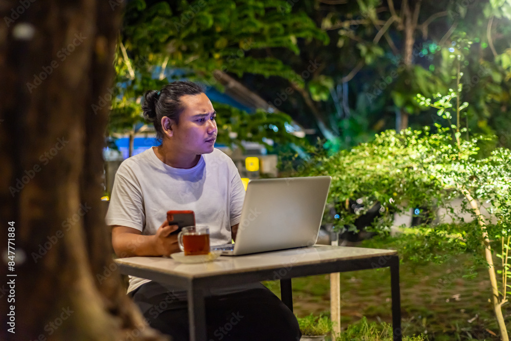 Young Asian man, digital nomad working remotely from cafe, using laptop and smartphone. young Asian men are hanging out in a cafe with a nature theme at night