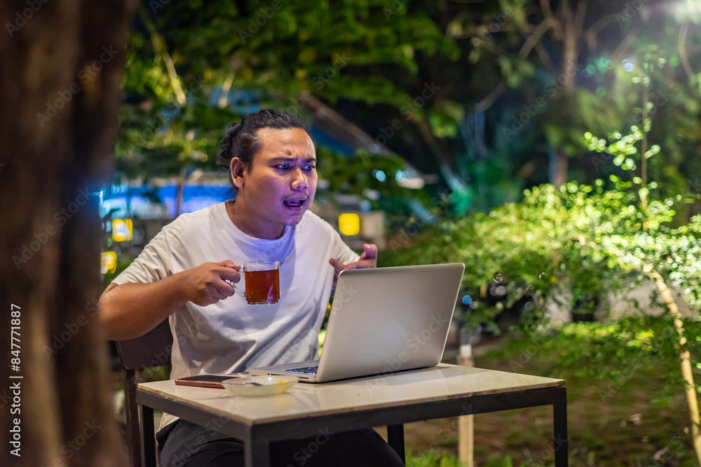 Young Asian man, digital nomad working remotely from cafe, using laptop and smartphone. young Asian men are hanging out in a cafe with a nature theme at night