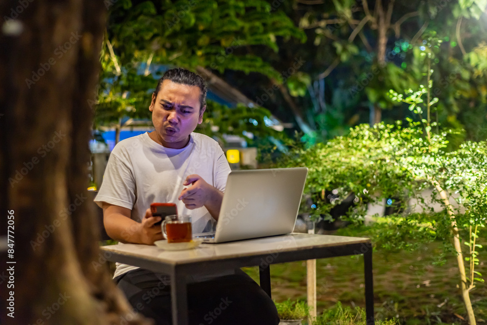 Young Asian man, digital nomad working remotely from cafe, using laptop and smartphone. young Asian men are hanging out in a cafe with a nature theme at night