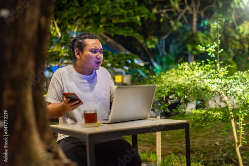 Obraz na płótnie Young Asian man, digital nomad working remotely from cafe, using laptop and smartphone. young Asian men are hanging out in a cafe with a nature theme at night w salonie
