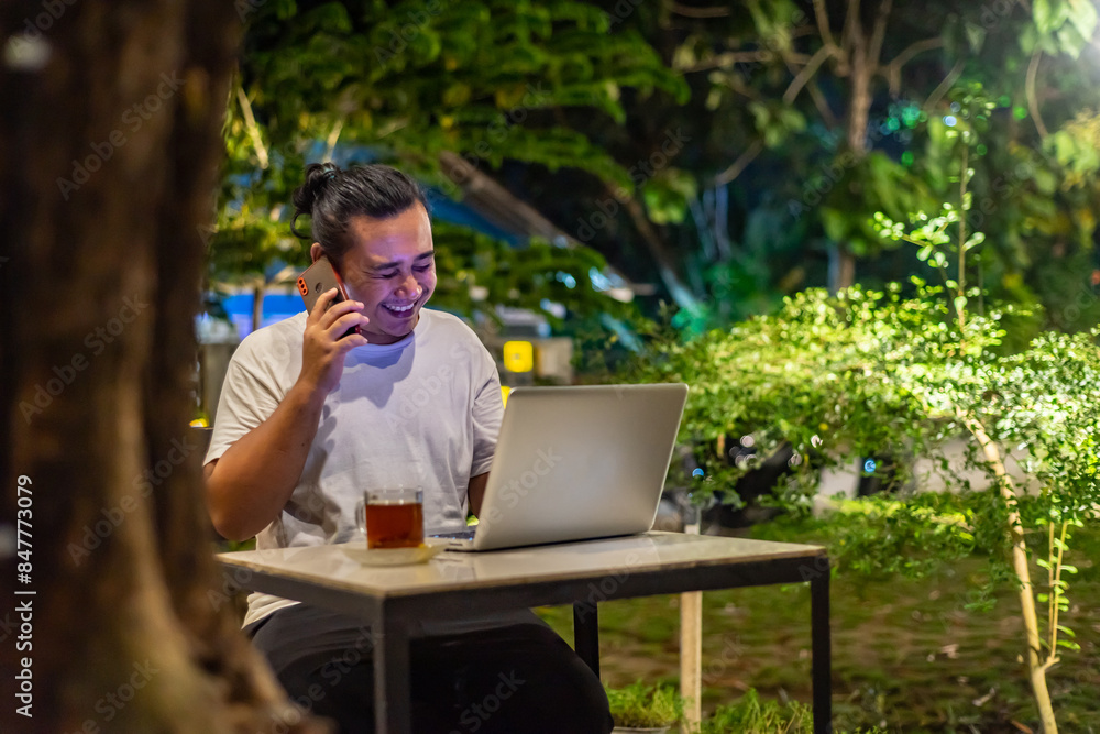 Young Asian man, digital nomad working remotely from cafe, using laptop and smartphone. young Asian men are hanging out in a cafe with a nature theme at night