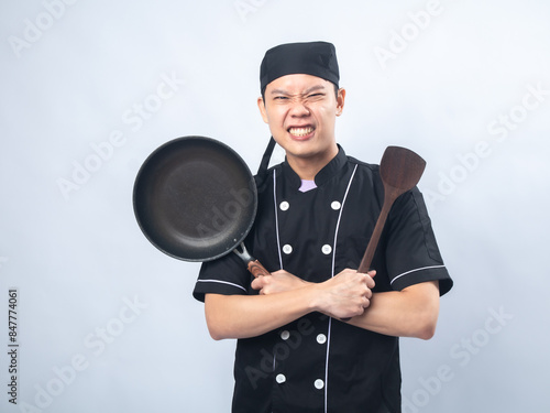 Smiling male chef in a black uniform and chef hat, holding a frying pan and a wooden spatula against a plain light gray background. The chef conveys enthusiasm and professionalism photo