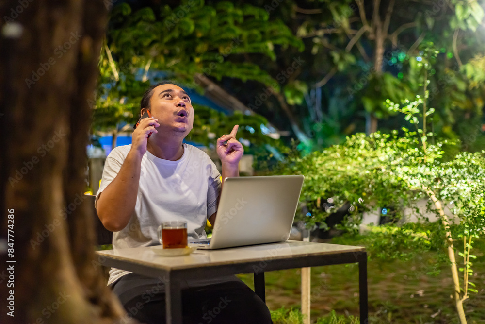 Young Asian man, digital nomad working remotely from cafe, using laptop and smartphone. young Asian men are hanging out in a cafe with a nature theme at night