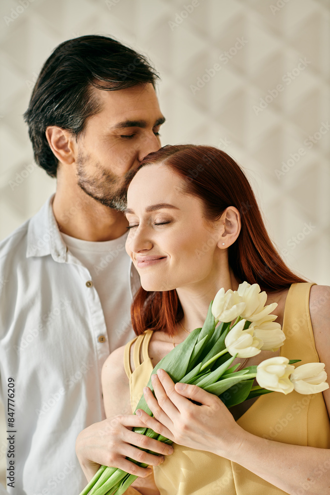 Obraz premium A redhead woman and bearded man embrace and kiss while holding a bouquet of tulips in a modern apartment.