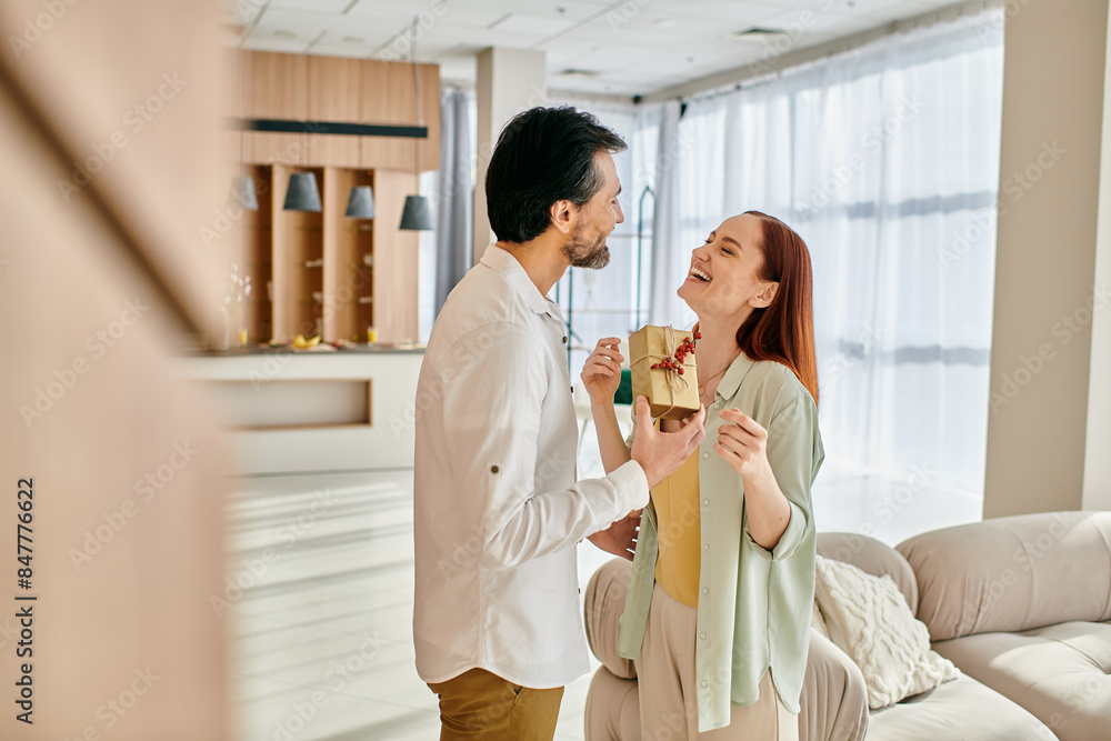 A redhead woman and a bearded man are standing in a modern living room, joyfully holding a gift between them. A redhead woman and a bearded man are standing in a modern living room, joyfully holding a gift between them.