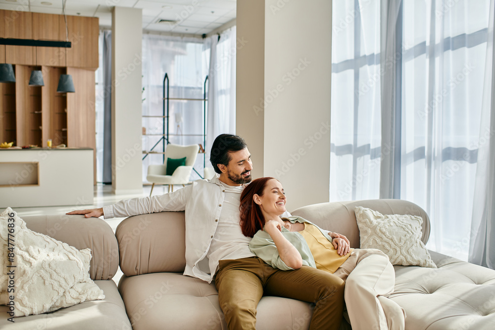 A man and woman, a redhead and a bearded man, relax on a couch in a modern living room, enjoying quality time together. A man and woman, a redhead and a bearded man, relax on a couch in a modern living room, enjoying quality time together.