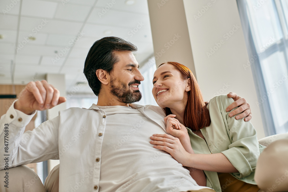 A beautiful adult couple, a redhead woman and a bearded man, sit together on a modern sofa in their apartment, enjoying quality time. A beautiful adult couple, a redhead woman and a bearded man, sit together on a modern sofa in their apartment, enjoying quality time.