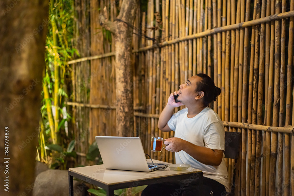 Young Asian man, digital nomad working remotely from cafe, using laptop and smartphone. young Asian men are hanging out in a cafe with a nature theme at night