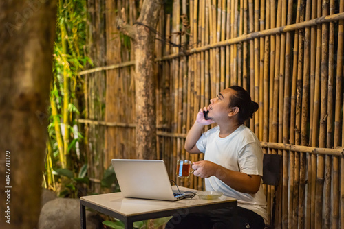 Obraz na płótnie Young Asian man, digital nomad working remotely from cafe, using laptop and smartphone. young Asian men are hanging out in a cafe with a nature theme at night w salonie