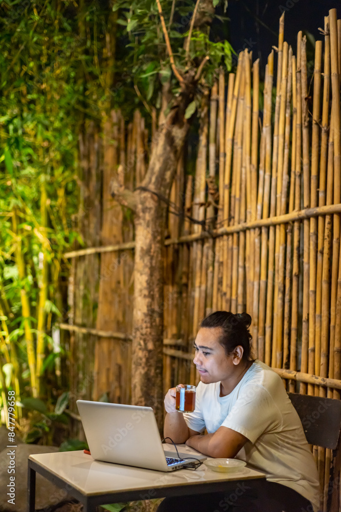 Young Asian man, digital nomad working remotely from cafe, using laptop and smartphone. young Asian men are hanging out in a cafe with a nature theme at night