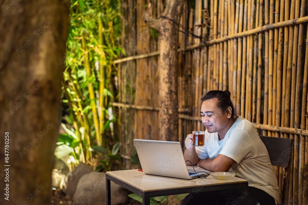 Young Asian man, digital nomad working remotely from cafe, using laptop and smartphone. young Asian men are hanging out in a cafe with a nature theme at night