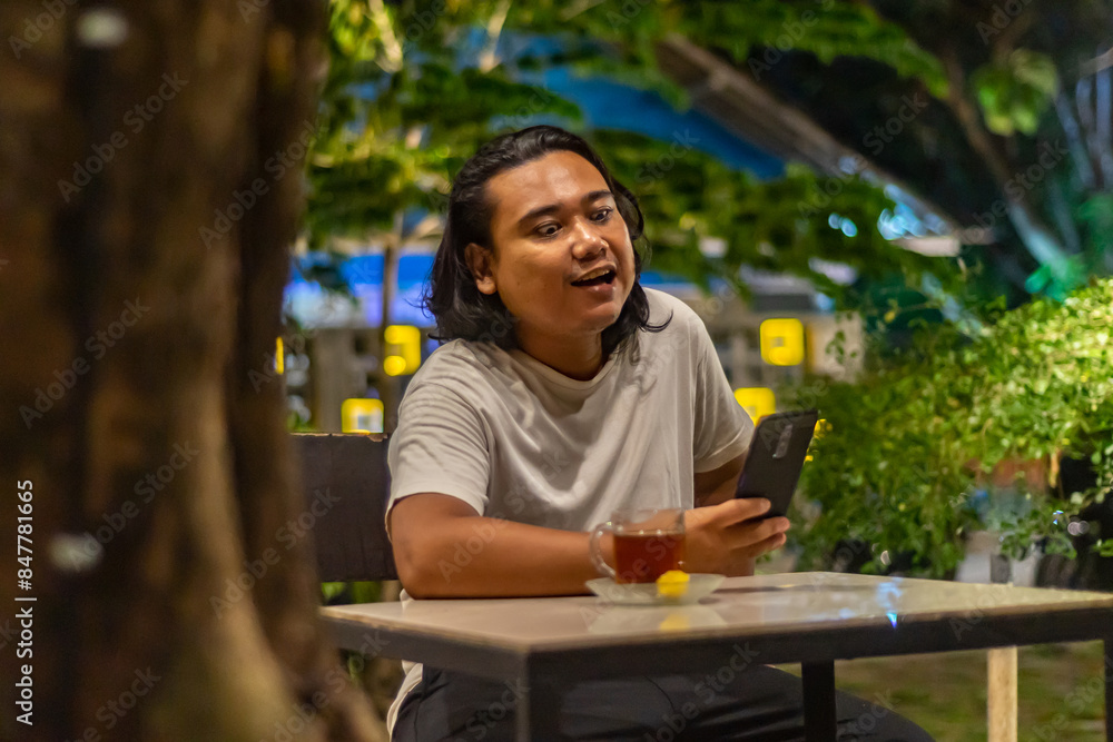 Young Asian man, digital nomad working remotely from cafe, using laptop and smartphone. young Asian men are hanging out in a cafe with a nature theme at night