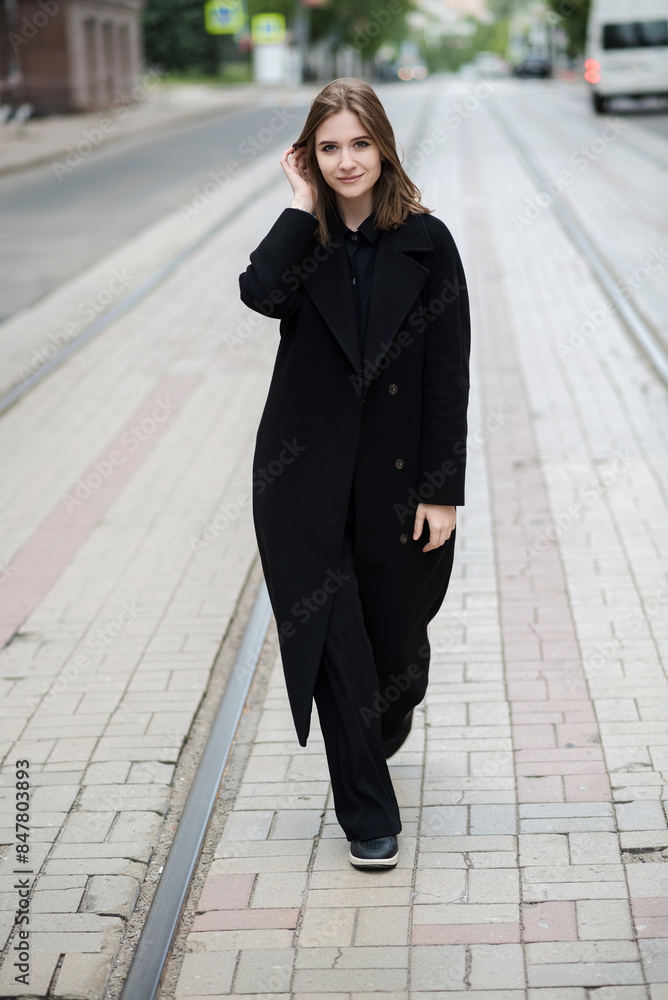 Attractive caucasian young woman in black coat walking down city street on warm autumn day. Outdoors portrait. Model in stylish clothes. Lifestyle, fashion concept. Attractive caucasian young woman in black coat walking down city street on warm autumn day. Outdoors portrait. Model in stylish clothes. Lifestyle, fashion concept.